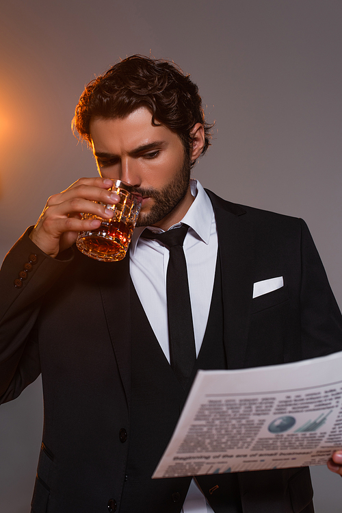 serious man in black blazer reading newspaper and drinking whiskey on grey background with light