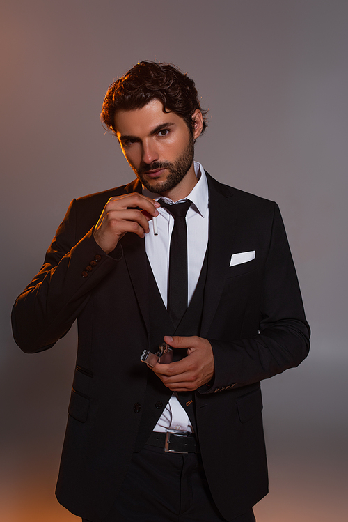 bearded man in black suit looking at camera while holding cigarette and lighter on grey background