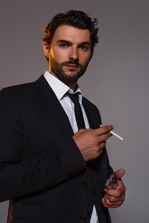 stylish man in black suit looking at camera while holding cigarette and lighter isolated on grey