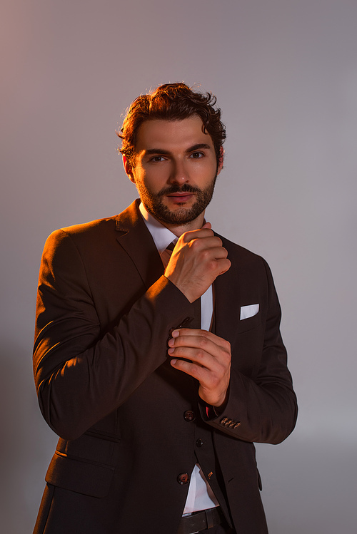 elegant man buttoning sleeve of black blazer while looking at camera isolated on grey