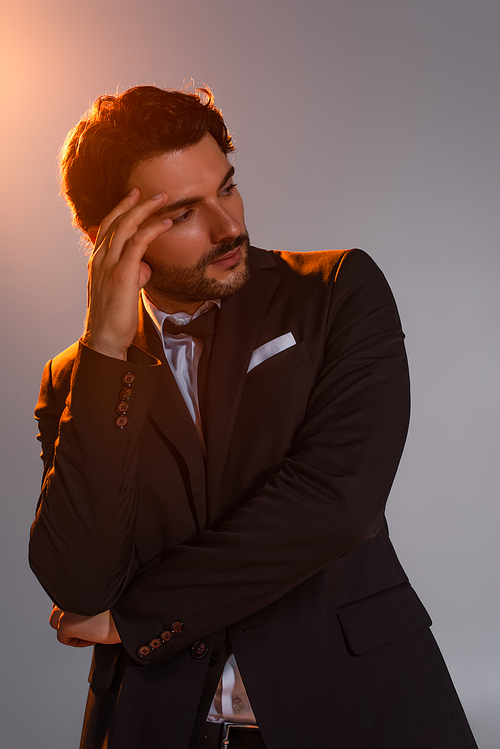 stylish man holding hand near face and looking away on grey background with light