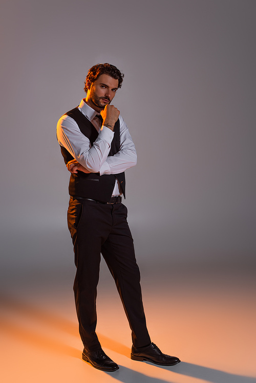 full length view of elegant brunette man looking at camera while standing on grey background with light