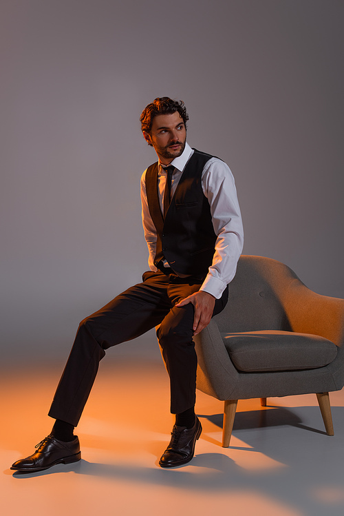 full length view of elegant businessman looking away near armchair on grey background with light