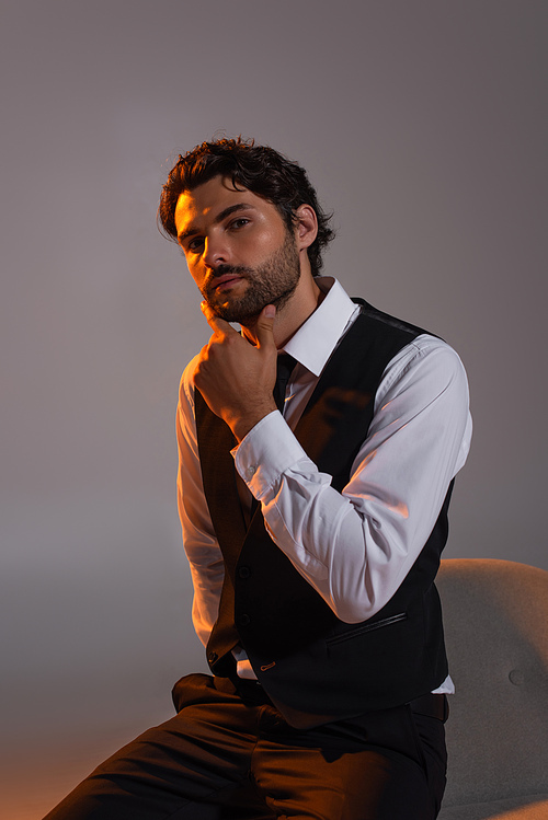 stylish brunette man in white shirt and black vest touching chin and looking at camera on grey background