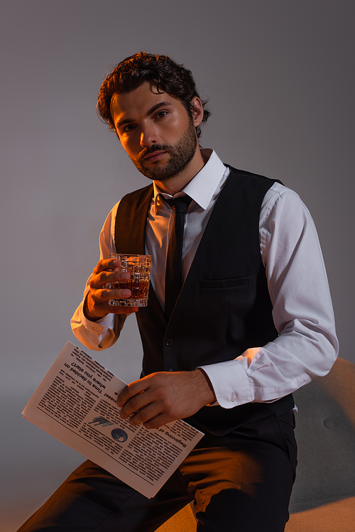 stylish man with glass of whiskey and newspaper looking at camera on grey background