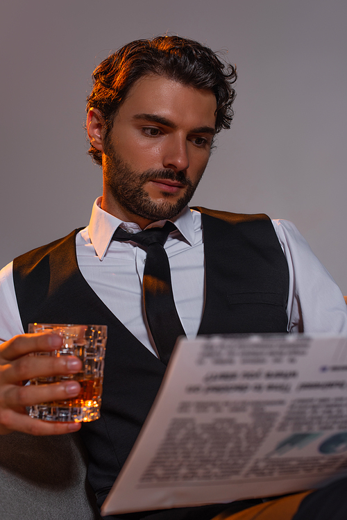 brunette businessman holding glass of whiskey and reading newspaper isolated on grey