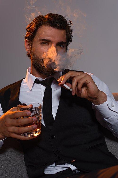 stylish man with glass of whiskey smoking cigarette and looking at camera on grey background