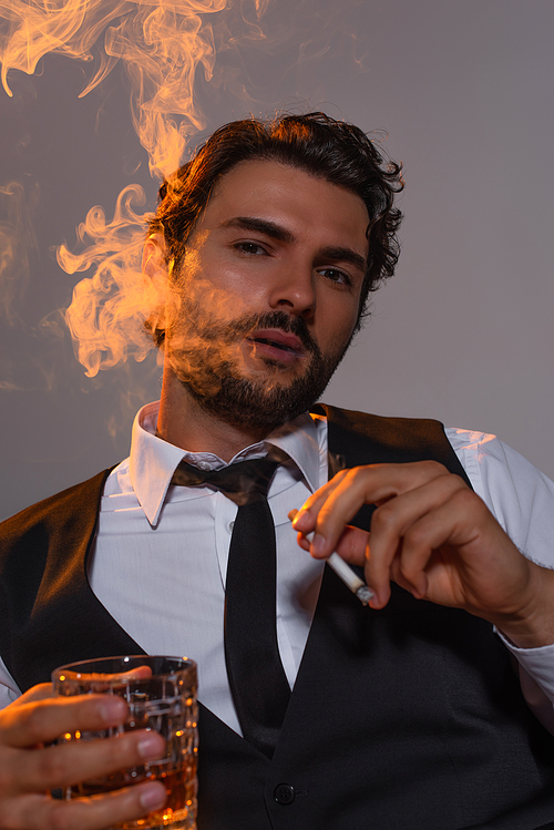 stylish man with glass of whiskey looking at camera and smoking on grey background with light