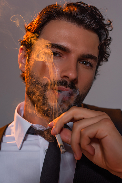 close up view of stylish man looking a camera through cigarette smoke on grey background