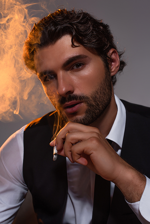 bearded businessman smoking and looking at camera on grey background with light