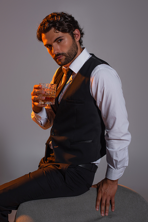 man in black vest and white shirt looking at camera while holding glass of whiskey isolated on grey