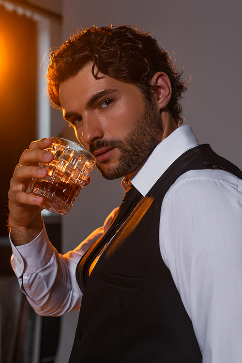 brunette man in white shirt and black vest holding glass of whiskey on grey background with light