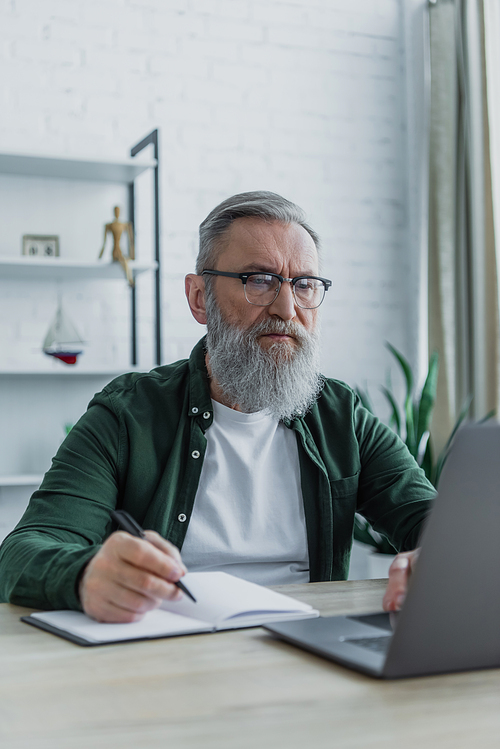 bearded senior man holding pen and using laptop while working from home