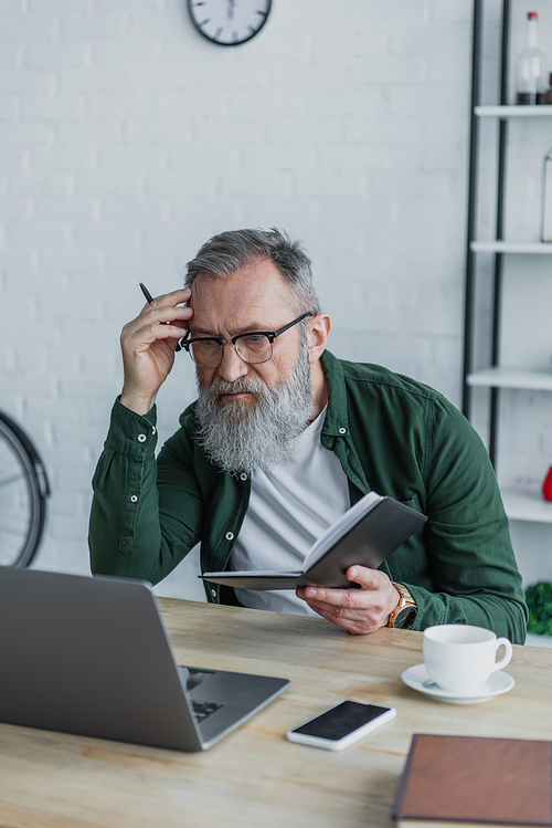 pensive and bearded senior man in eyeglasses holding pen and using laptop while working from home