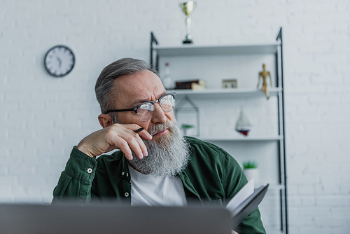 pensive and bearded senior man in eyeglasses holding pen and looking away while working from home