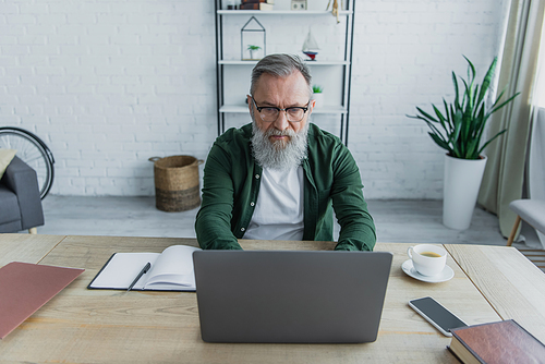 pensive and bearded senior man in eyeglasses using laptop while working from home