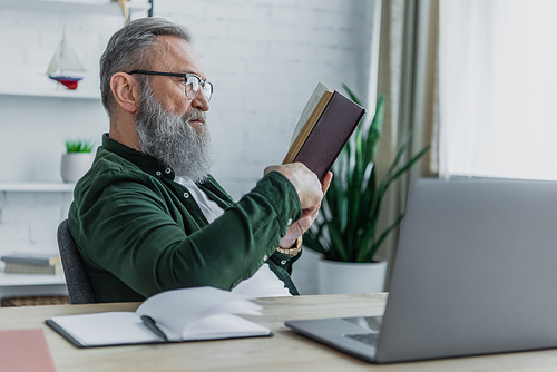 bearded senior man in eyeglasses reading book near laptop on desk