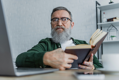 bearded senior man in eyeglasses looking at camera near laptop while holding book