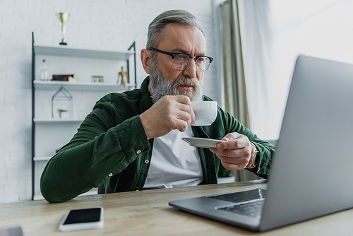 bearded senior man in eyeglasses drinking coffee and looking at laptop while working from home
