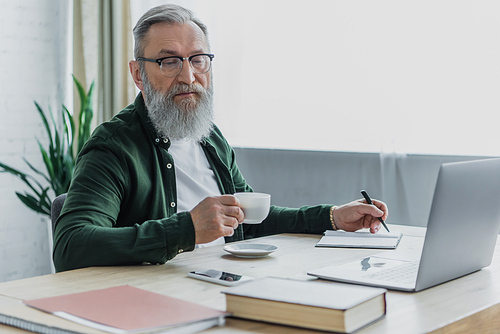 bearded senior man in eyeglasses holding cup of coffee and pen near laptop while working from home
