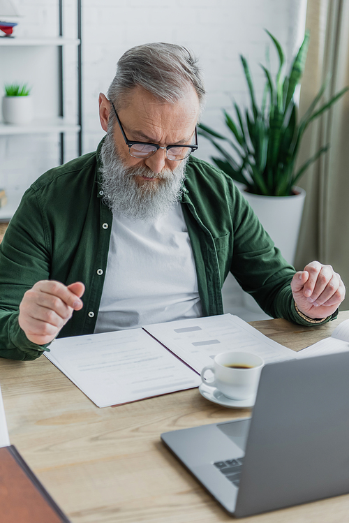 bearded senior man in eyeglasses looking at documents near cup of coffee and laptop while working from home