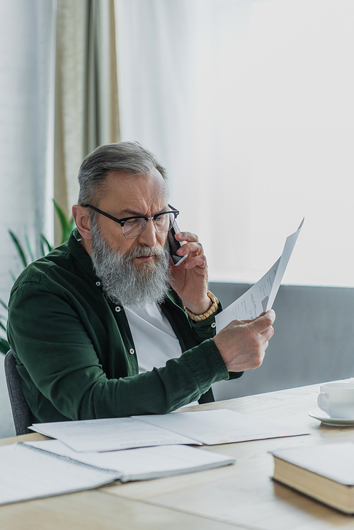 bearded senior man in eyeglasses talking on smartphone and holding document