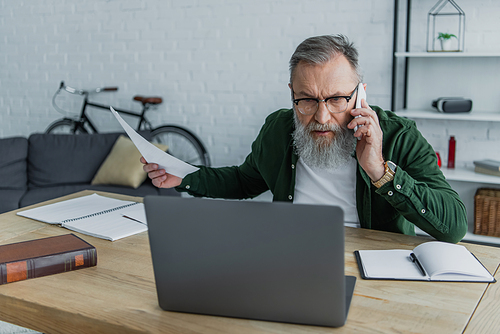 displeased senior man in eyeglasses talking on smartphone and holding document near laptop on desk