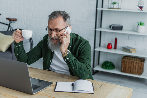 bearded senior man in eyeglasses holding cup and talking on smartphone near laptop