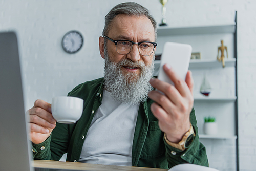 bearded senior man in eyeglasses holding cup and looking at smartphone