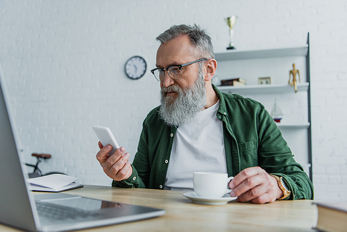bearded senior man in eyeglasses holding cup and looking at smartphone near laptop on desk