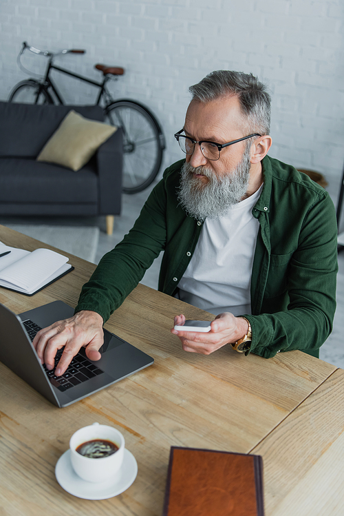 bearded senior man in eyeglasses holding smartphone while using laptop at home