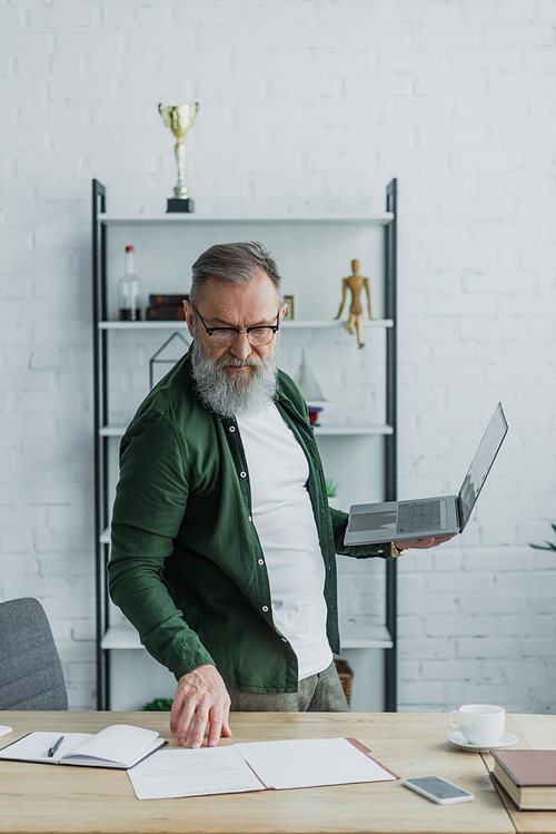 bearded senior man in eyeglasses standing with laptop while looking at papers on desk
