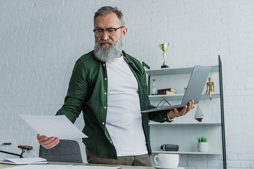 bearded senior man in eyeglasses standing with laptop while looking at papers at home