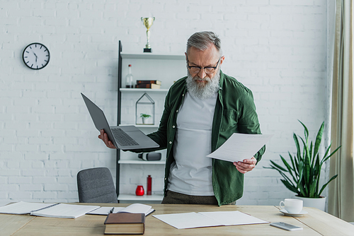 bearded senior man in eyeglasses standing with laptop while looking at documents on desk
