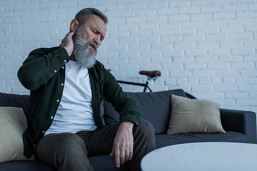 unhappy senior man with beard sitting on couch and touching neck while suffering from pain