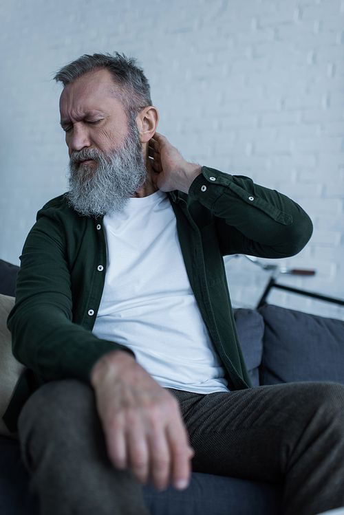 upset senior man with beard and closed eyes sitting on couch and having neck pain