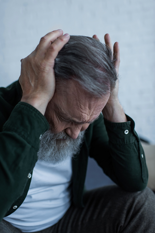stressed senior man with beard touching head while suffering from headache at home
