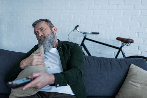 depressed senior man holding remote controller and pillow while watching tv in living room