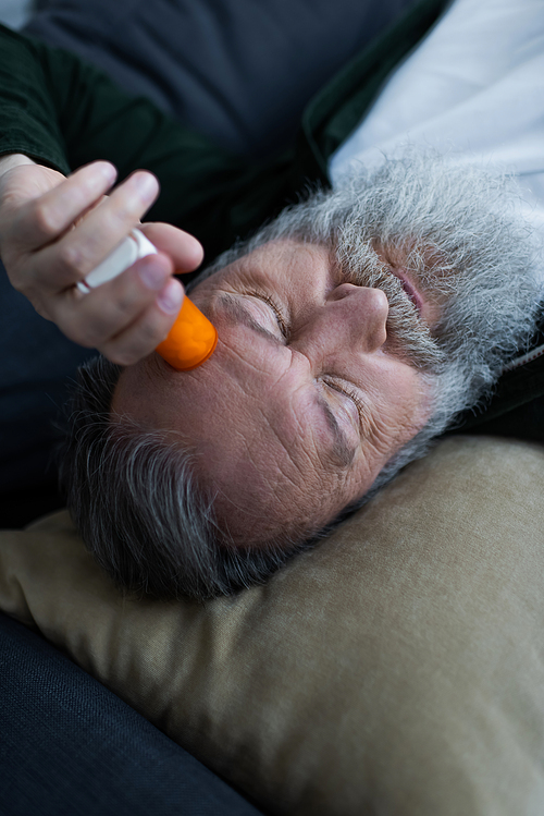unhappy senior man holding bottle with anti-depressant medication while lying on couch in living room