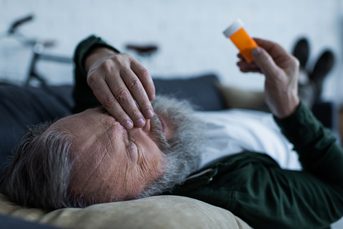 depressed senior man holding bottle with medication while lying on couch in living room