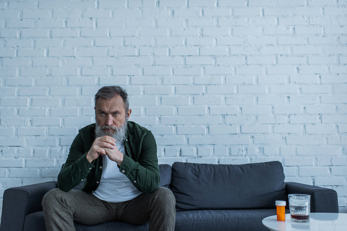 depressed senior man with grey hair sitting on couch near bottle with medication and glass of whiskey on coffee table