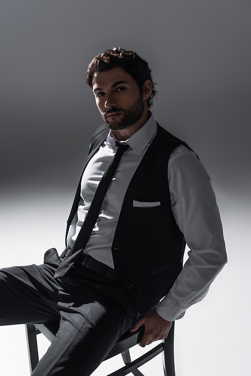 brunette man in elegant formal wear looking at camera while sitting on grey