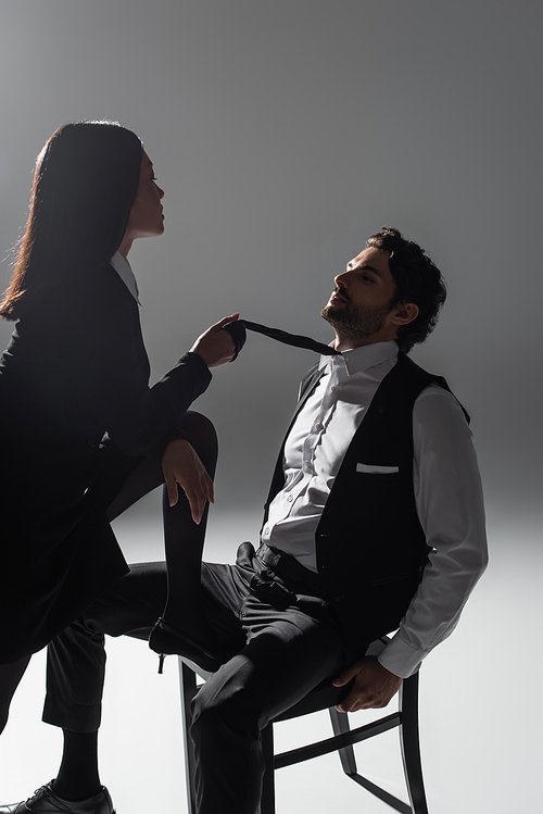 hot asian woman holding tie of elegant man sitting on chair on grey background