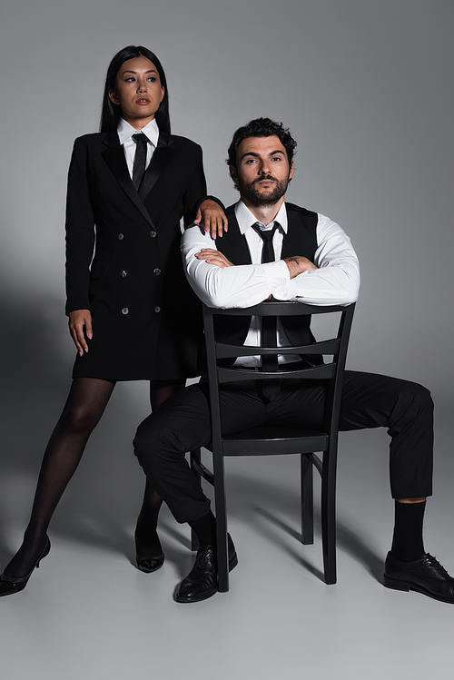 sexy asian woman in black blazer dress near elegant man sitting on chair on grey background