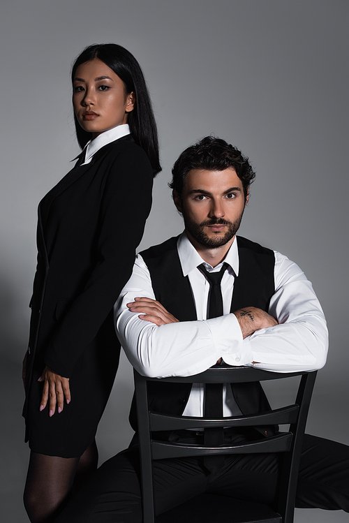 elegant man sitting on chair with crossed arms near sexy asian woman on grey background