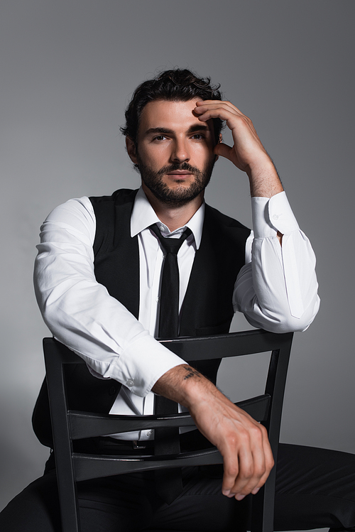 stylish man in black vest and tie sitting on chair and looking at camera isolated on grey