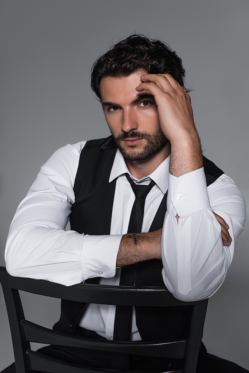 stylish man in black vest and tie looking at camera while sitting isolated on grey