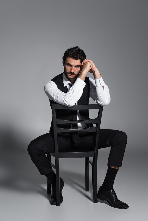 full length view of brunette man sitting on chair and looking at camera on grey