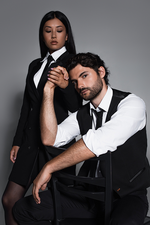 brunette man sitting on chair and holding hands with young asian woman on grey background