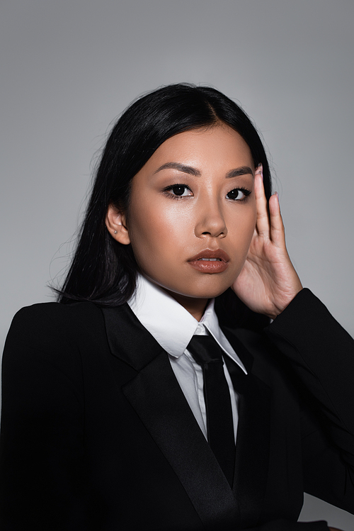 brunette asian woman in black blazer and tie looking at camera isolated on grey